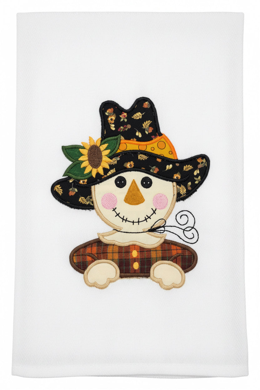 A white fabric tea towel featuring an embroidered autumn scarecrow design
