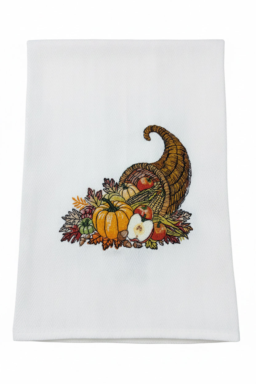 A white kitchen towel featuring a detailed embroidered autumn harvest design with a cornucopia, pumpkins, apples, and fall leaves.